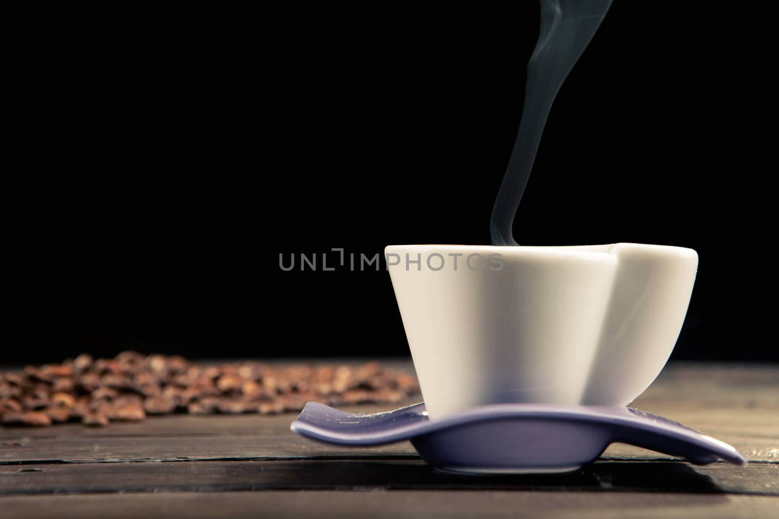 close up of a steaming cup of coffee with coffee beans scattered behind on a wooden table and a black background