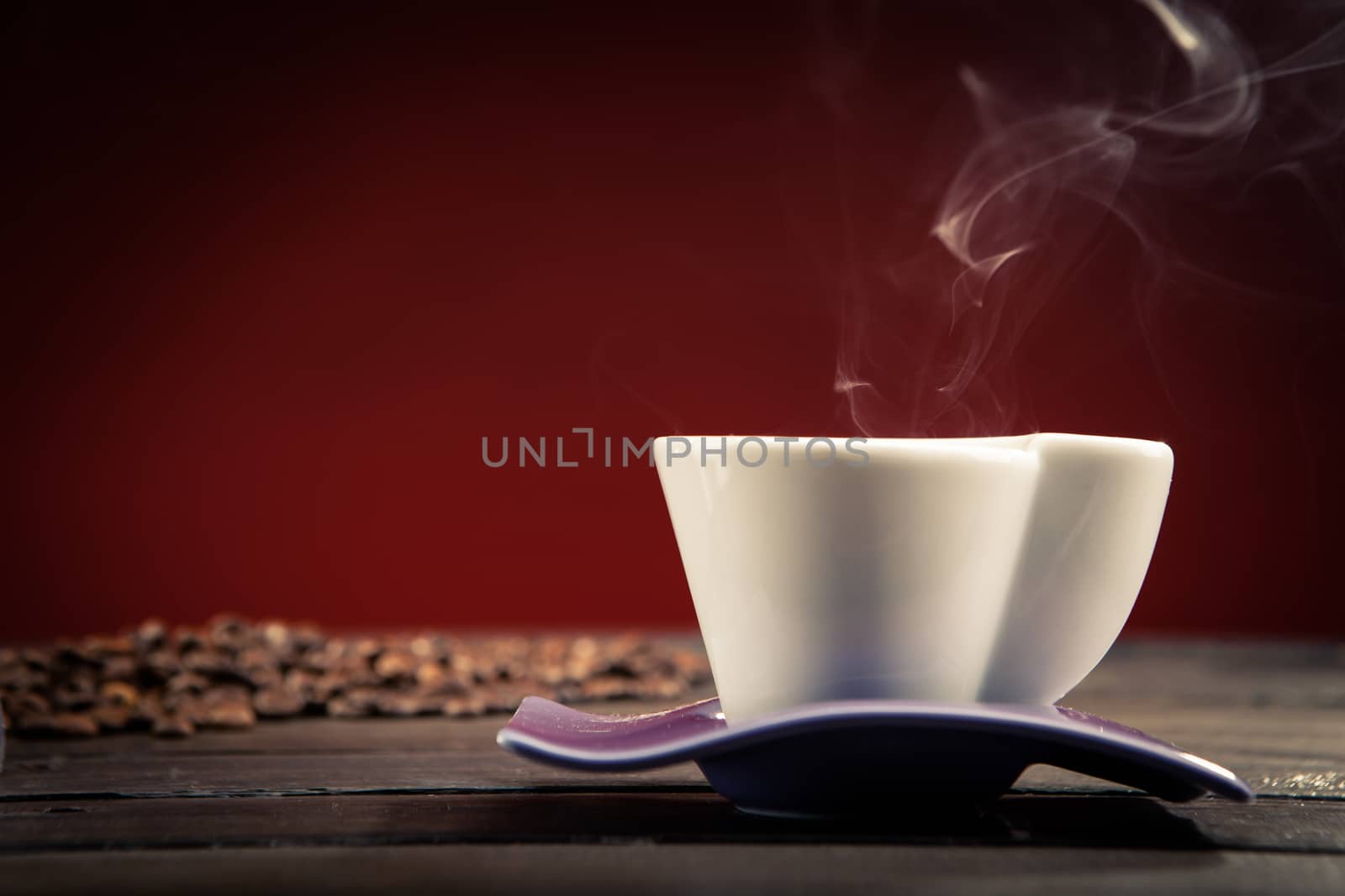 close up of a steaming cup of coffee with coffee beans scattered behind on a wooden table and a red background