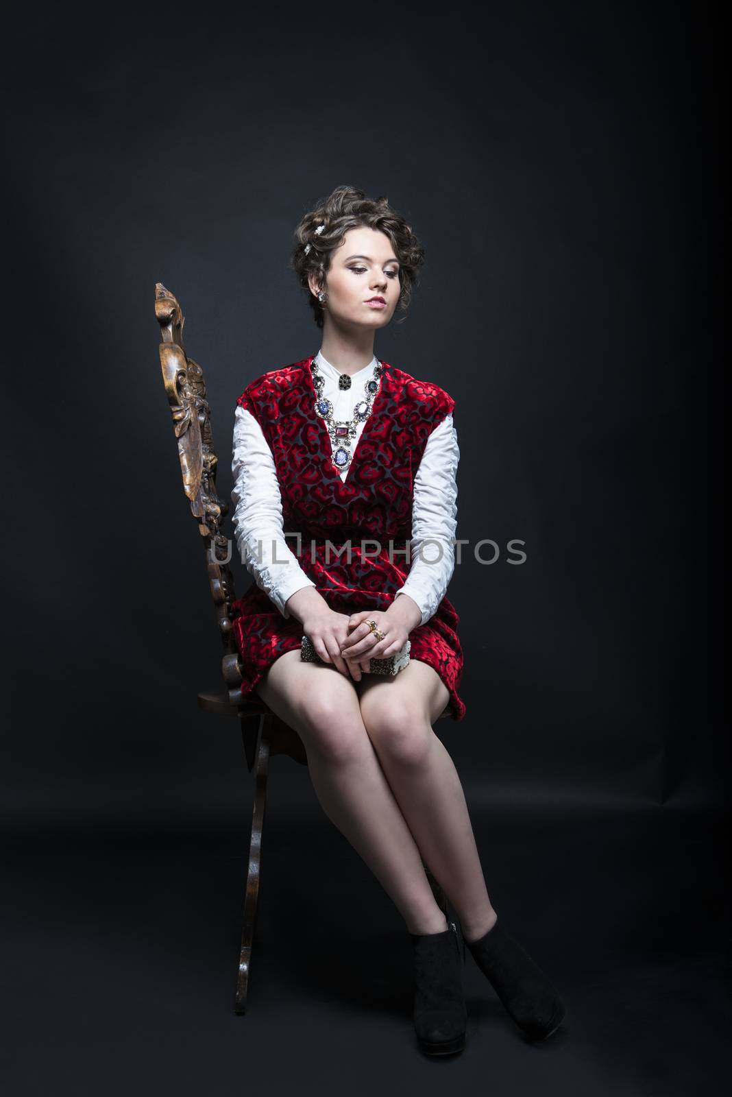 Portrait of beautiful Caucasian model in red Victorian-style velvet dress, seated on wooden chair