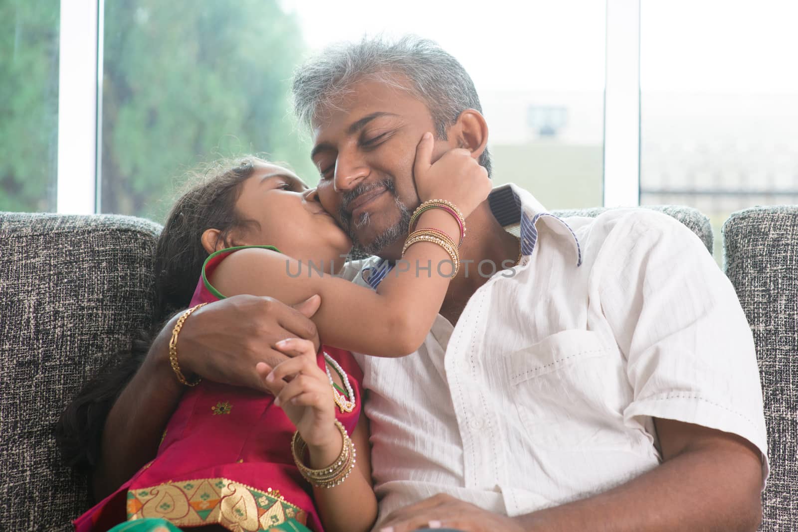 Happy Indian family at home. Asian sweet daughter kissing her father face indoor, sitting on sofa.