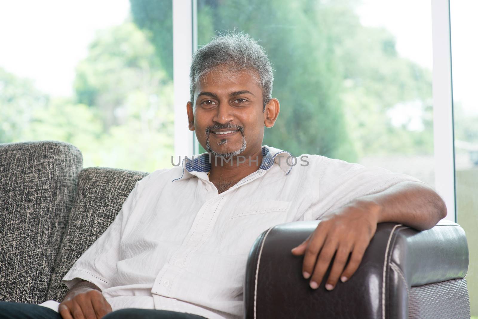 Portrait of mature Indian man sitting on sofa at home. Asian male relax on couch in house with interior.