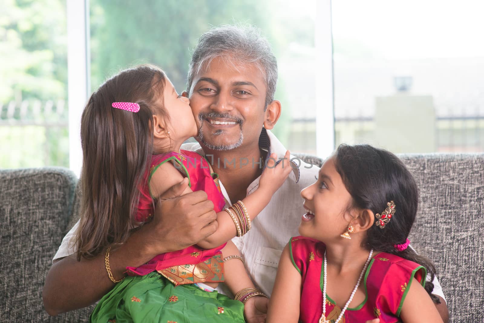 Happy Indian family at home. Asian girl kissing her father, sitting on sofa. Parent and children indoor lifestyle.