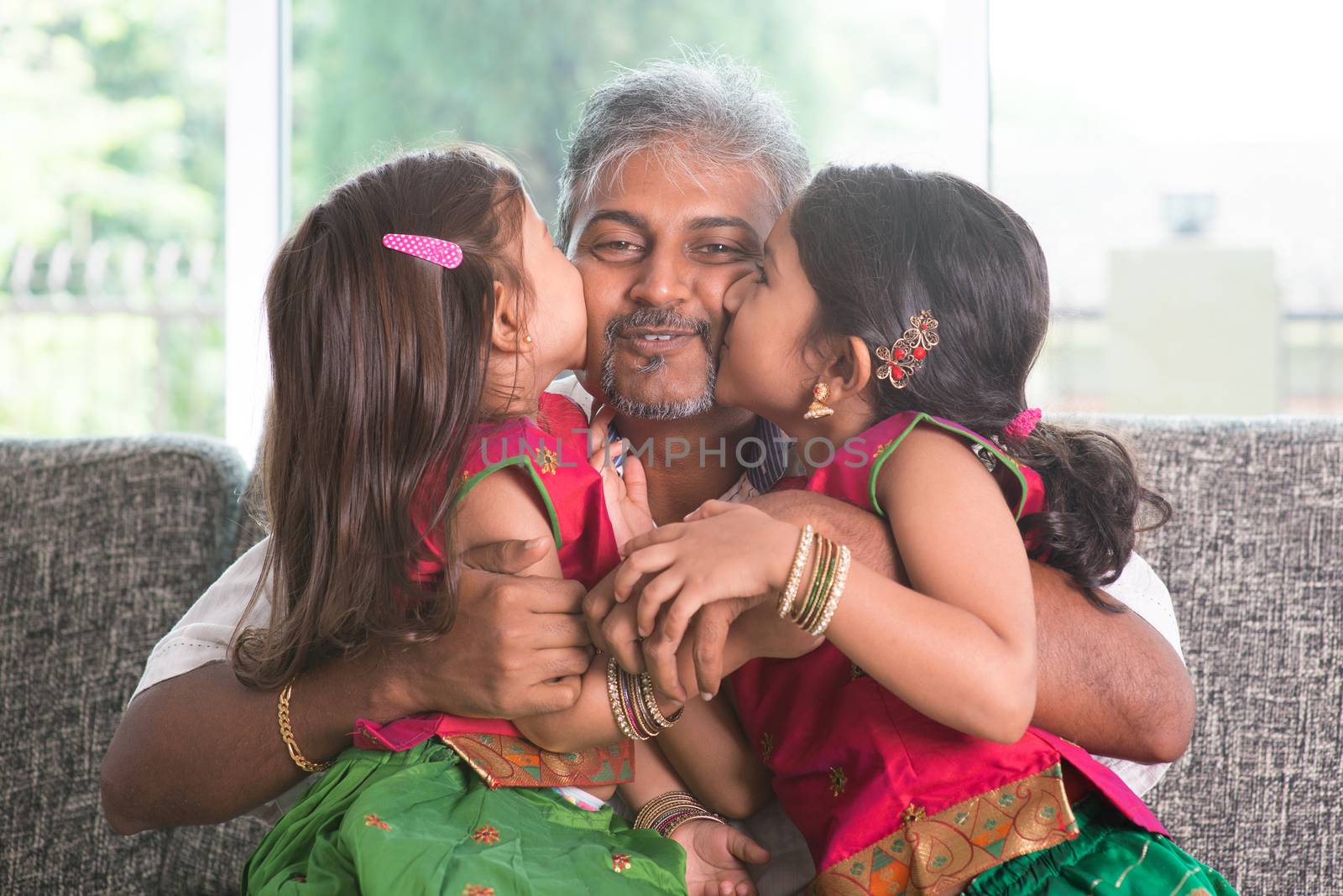 Happy Indian family at home. Two cute Asian girls kissing father, sitting on sofa. Parent and children indoor lifestyle.