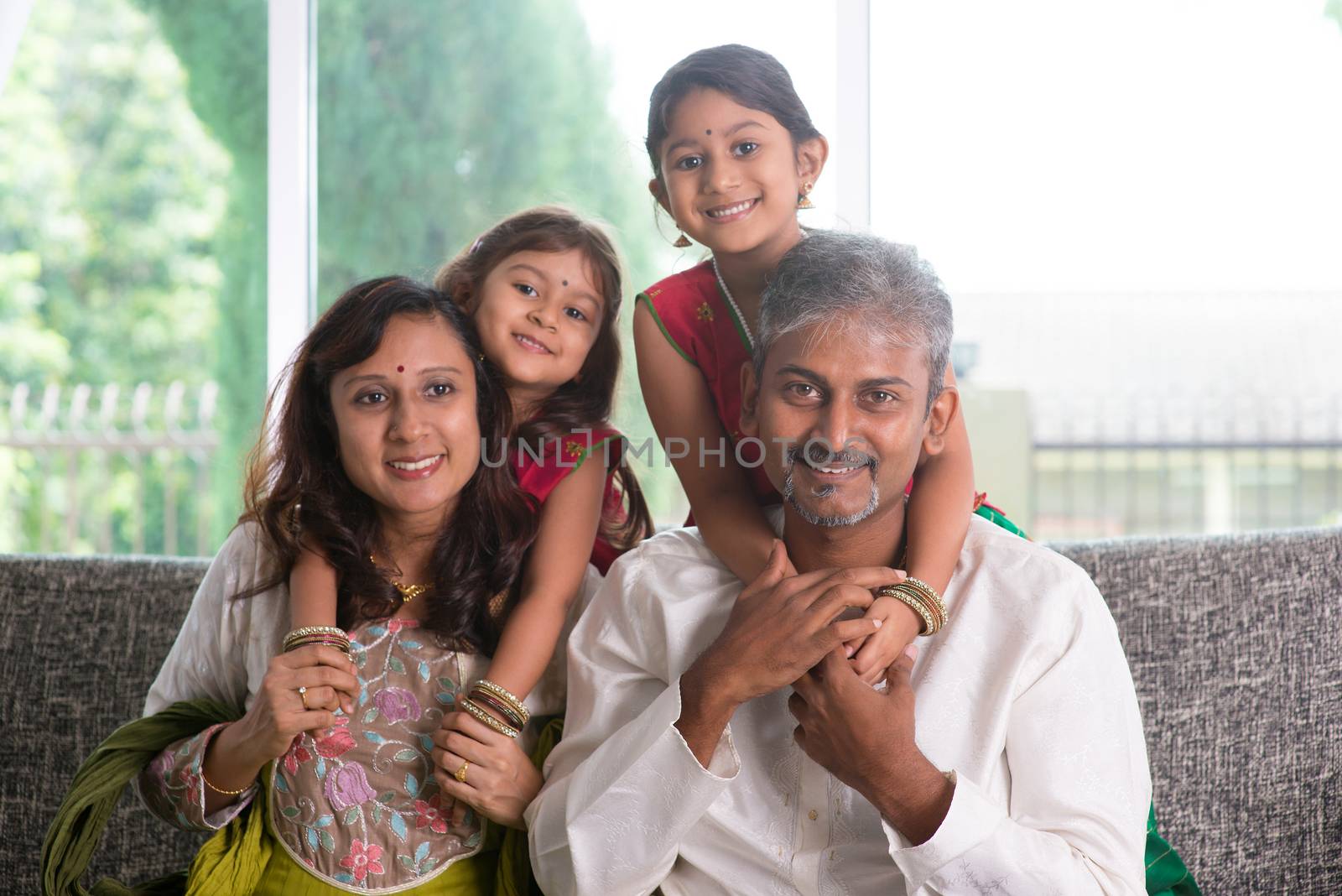 Happy Indian family at home. Asian parents piggyback their kids, sitting on sofa. Parents and children indoor lifestyle.