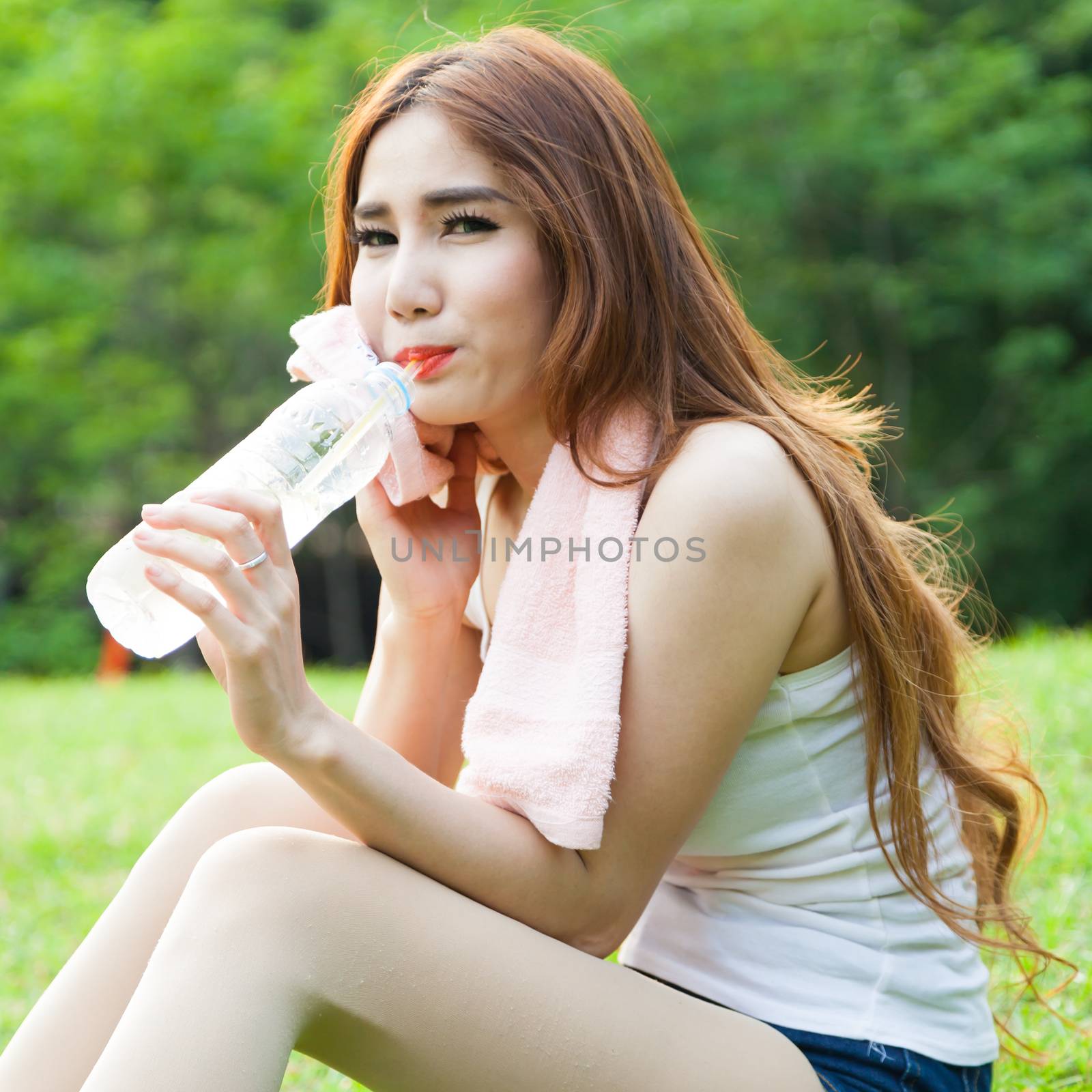 Woman sitting tired and drinking water after exercise. Within the lawn of park.