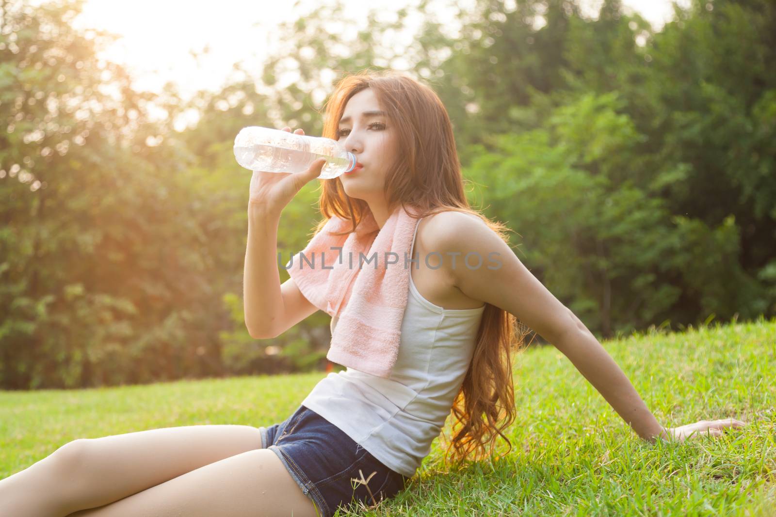 Woman sitting tired and drinking water after exercise. Within the lawn of park.