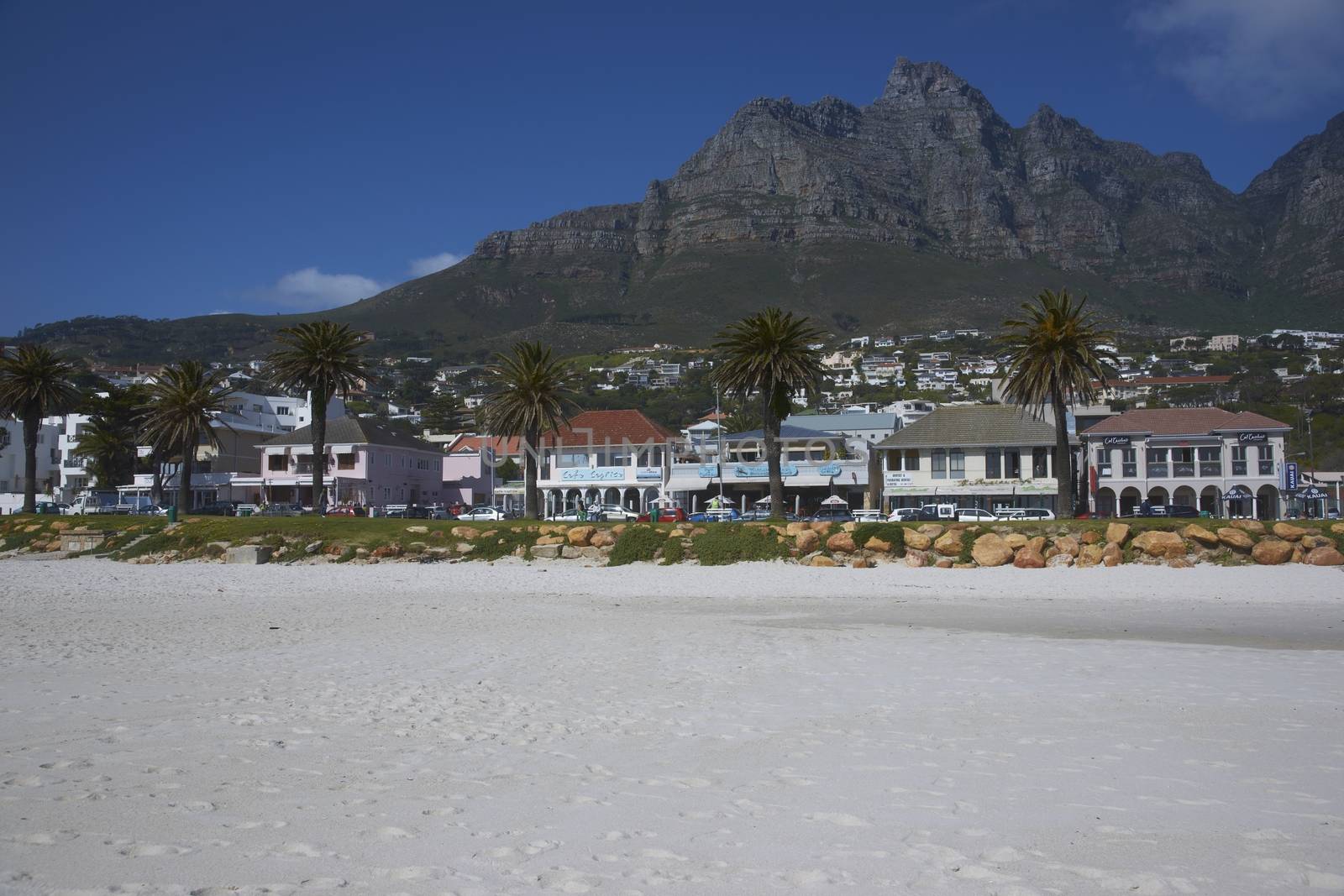 The Twelve Apostles mountains towering over the restaurants and bars lining the sandy beach at Camps Bay, Cape Town, South Africa