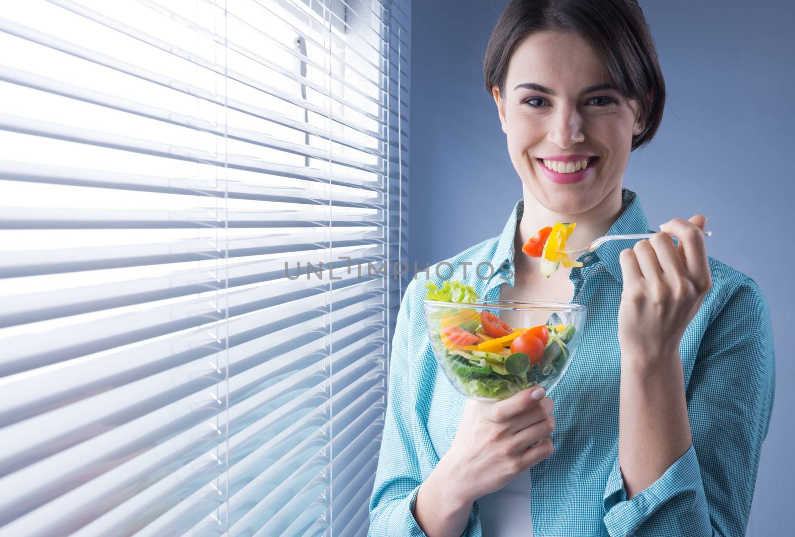Woman smiling at camera and eating salad in front of a window.