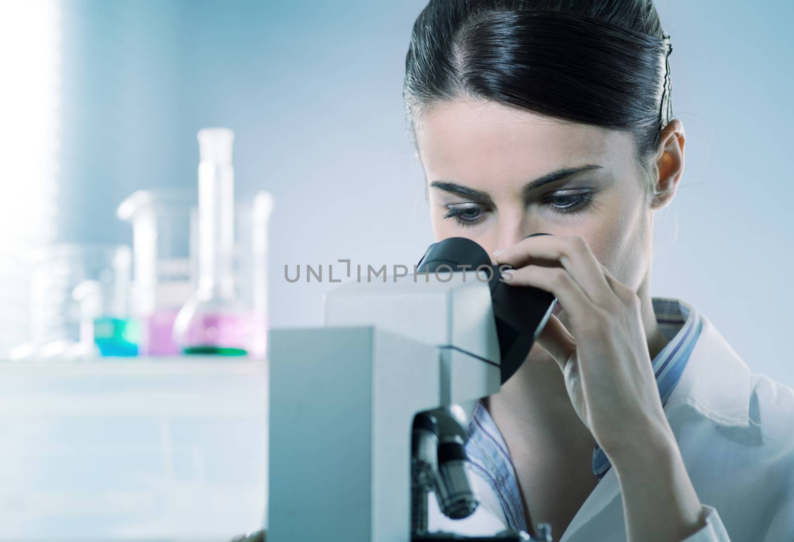 Young female researcher using microscope in the chemistry lab with laboratory glassware on background.