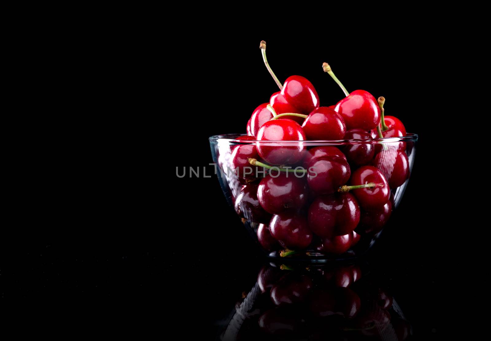 Juicy shiny cherries in a glass bowl on black background.