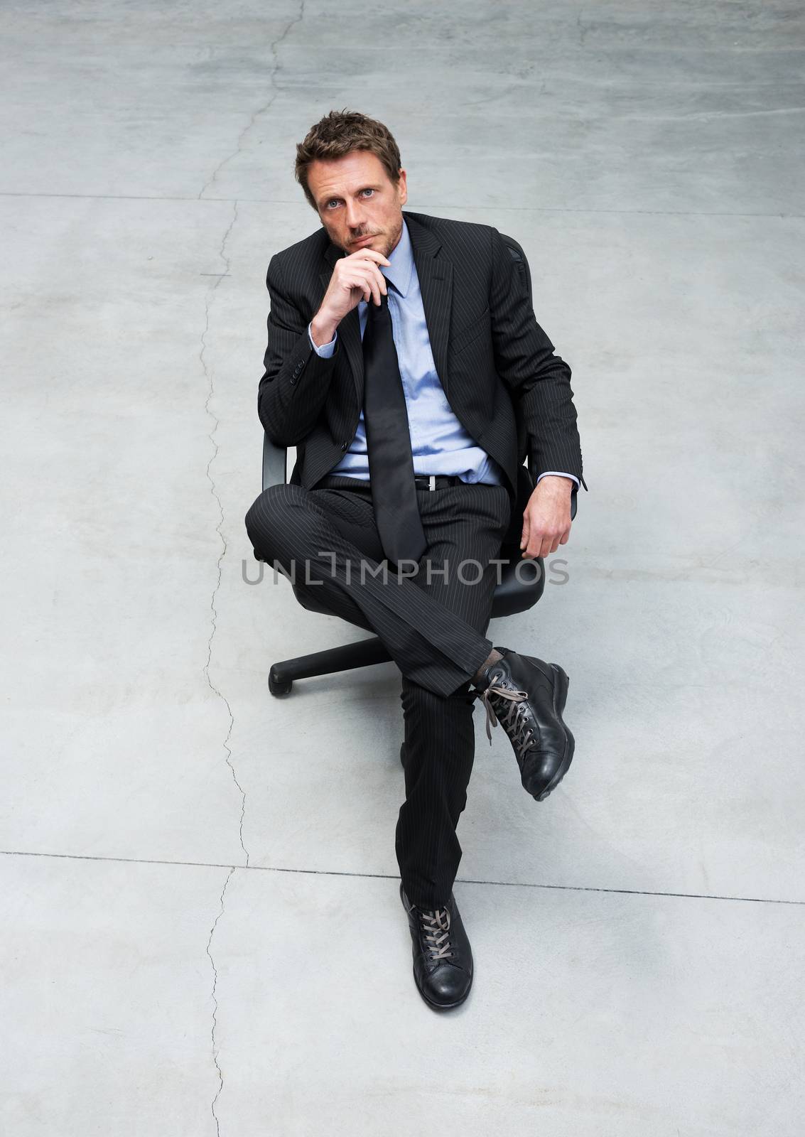 Smiling businessman sitting on an office chair with hand on chin against concrete floor background.