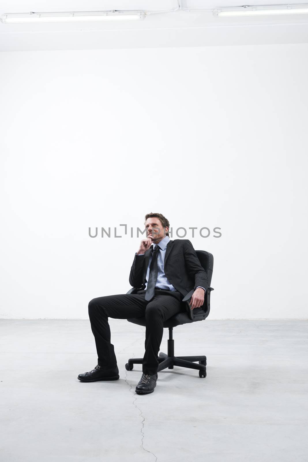 Businessman sitting in his new empty office looking around with hand on chin.