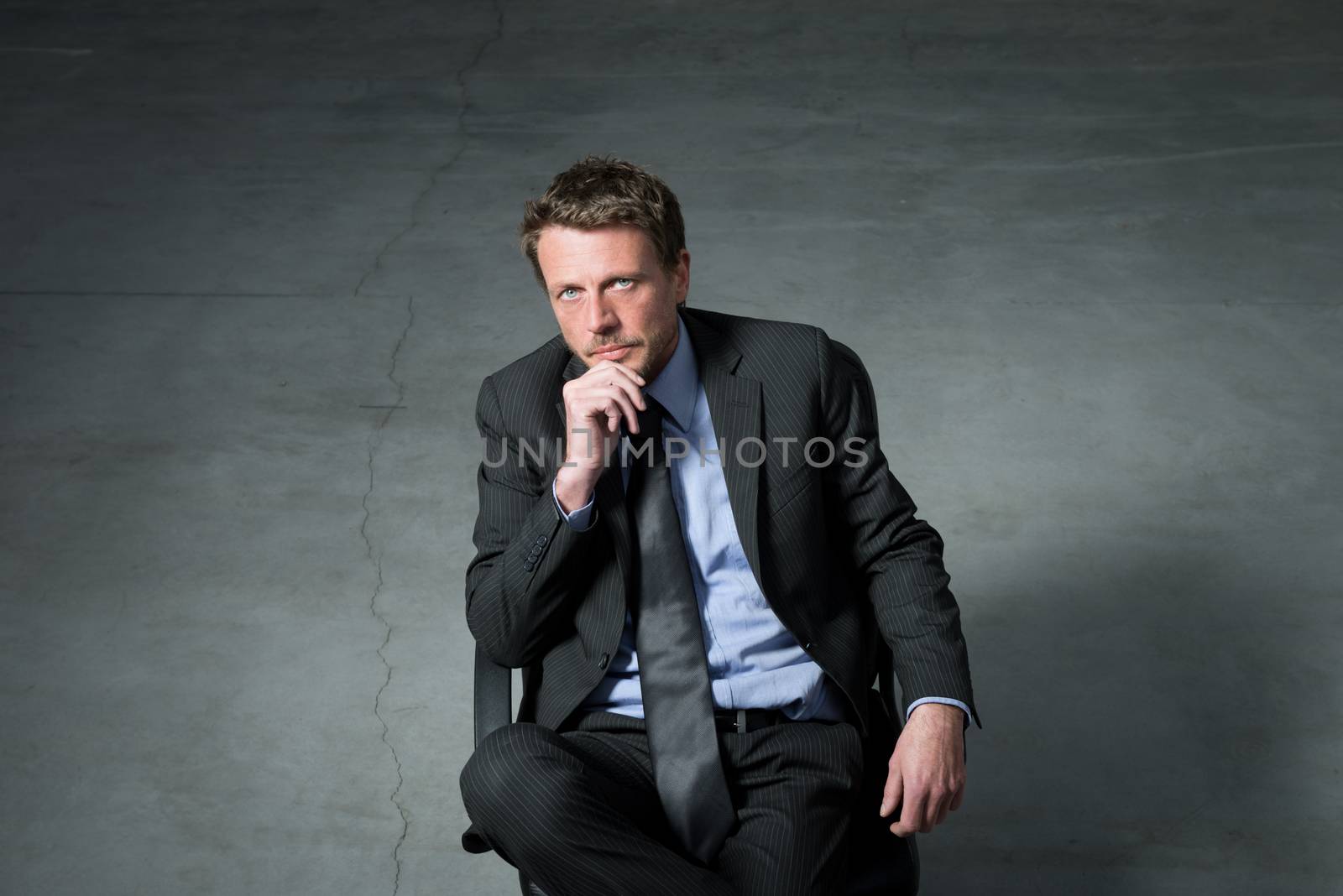 Businessman sitting on an office chair with hand on chin against concrete floor background.