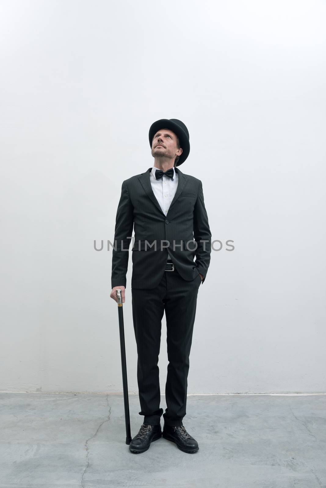 Classy gentleman standing on white background and concrete floor in elegant suit with cane and bowler hat, looking up.
