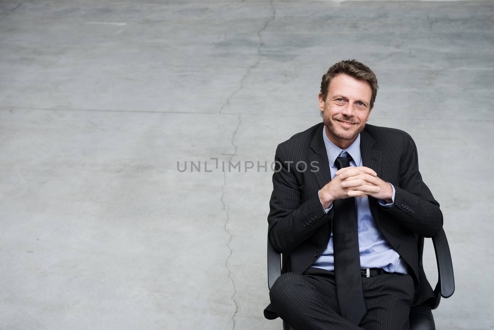 Smiling businessman sitting on office chair with hands clasped on concrete floor background.