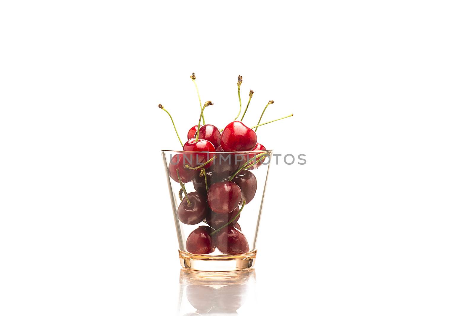 Horizontal close-up of a transparent glass full of red fresh cherries, natural source of vitamin C and minerals, with reflection on white