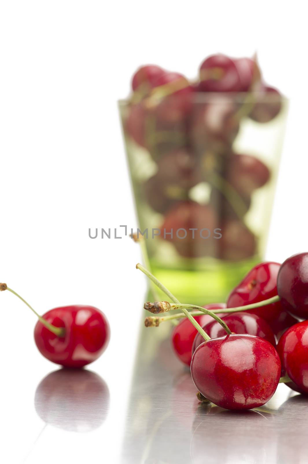 Loose delicious healthy ripe red cherries on a reflective surface with a glass full in the background with focus to the fruit in the front, on white with copyspace