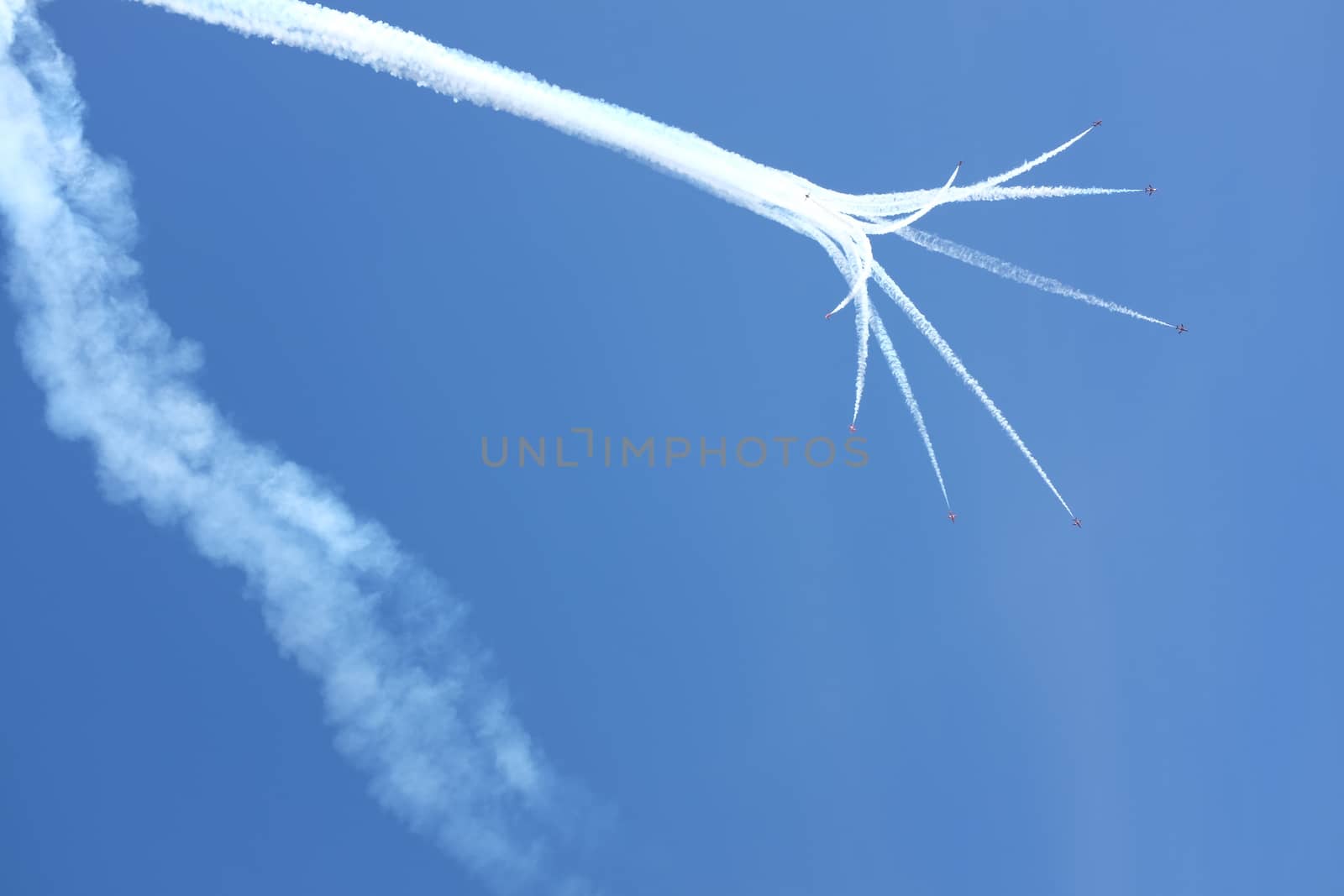 Vapour trails lead to a formation of red jet aircraft in display formation against a clear blue sky.