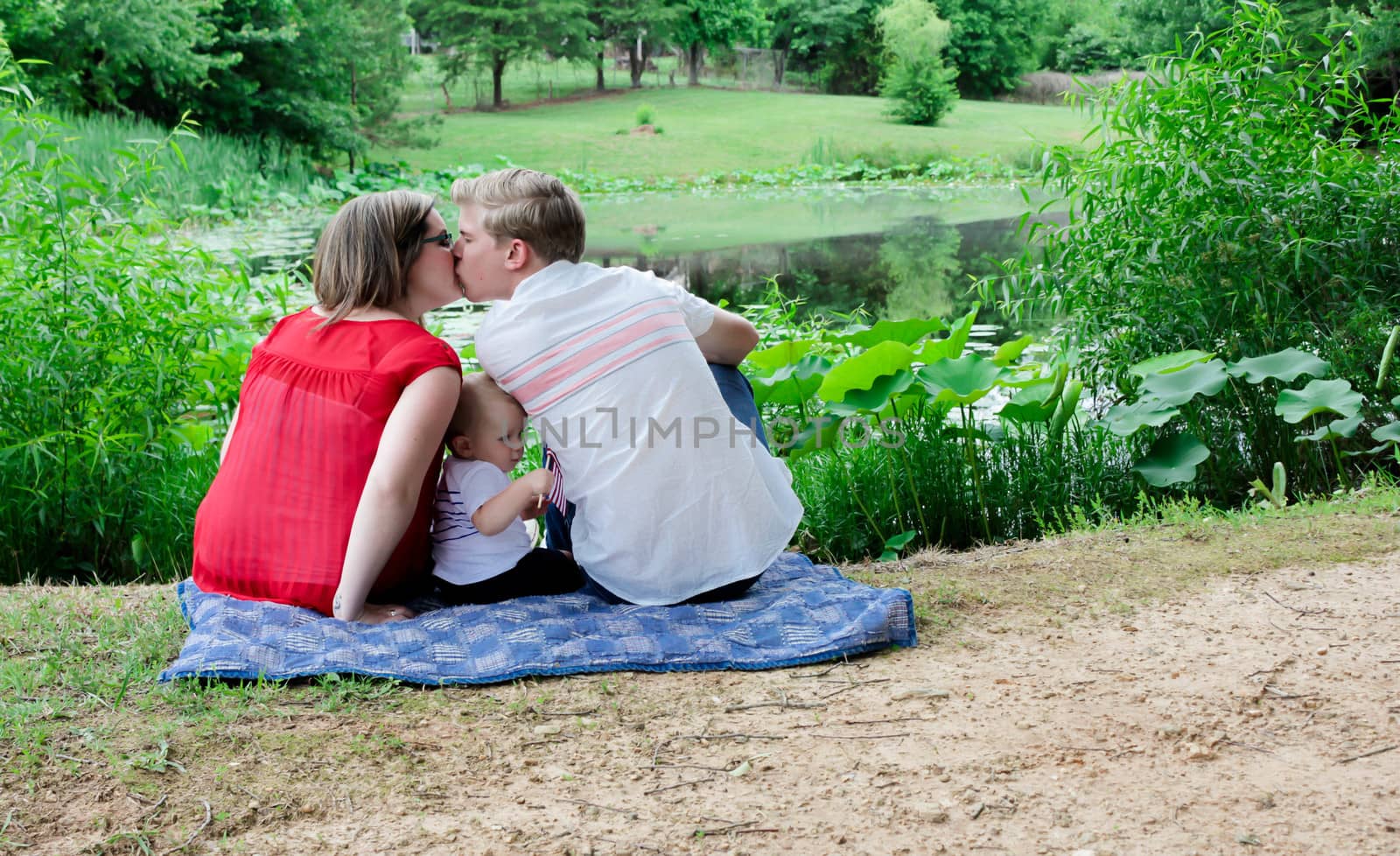 mother, father and baby sitting on a blanket by a pond.