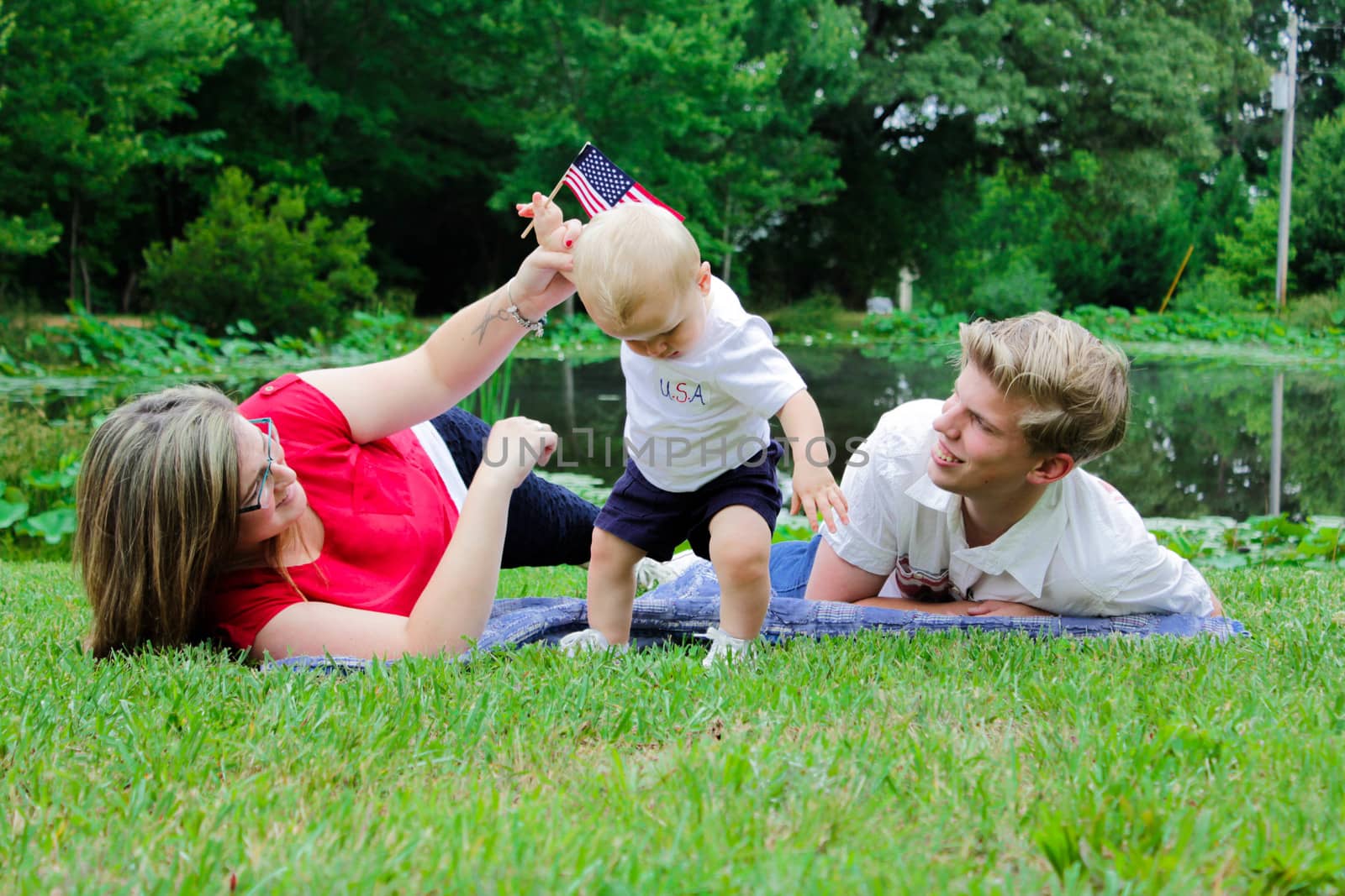 young mother and father laying on a blanket by a pool teaching their baby to stand on his own while he is holding an American flag