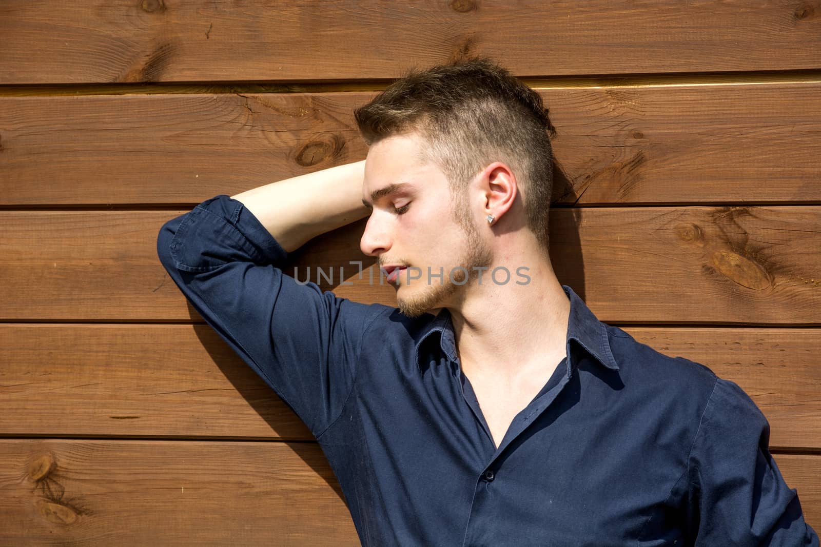 Handsome blond young man standing outside against wood wall with eyes closed