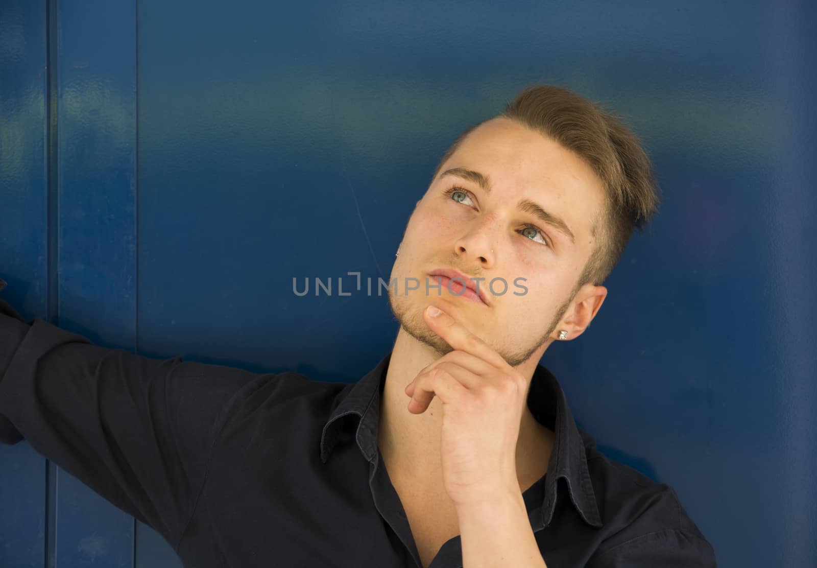 Attractive young man thinking, looking up with hand on his chin on blue background