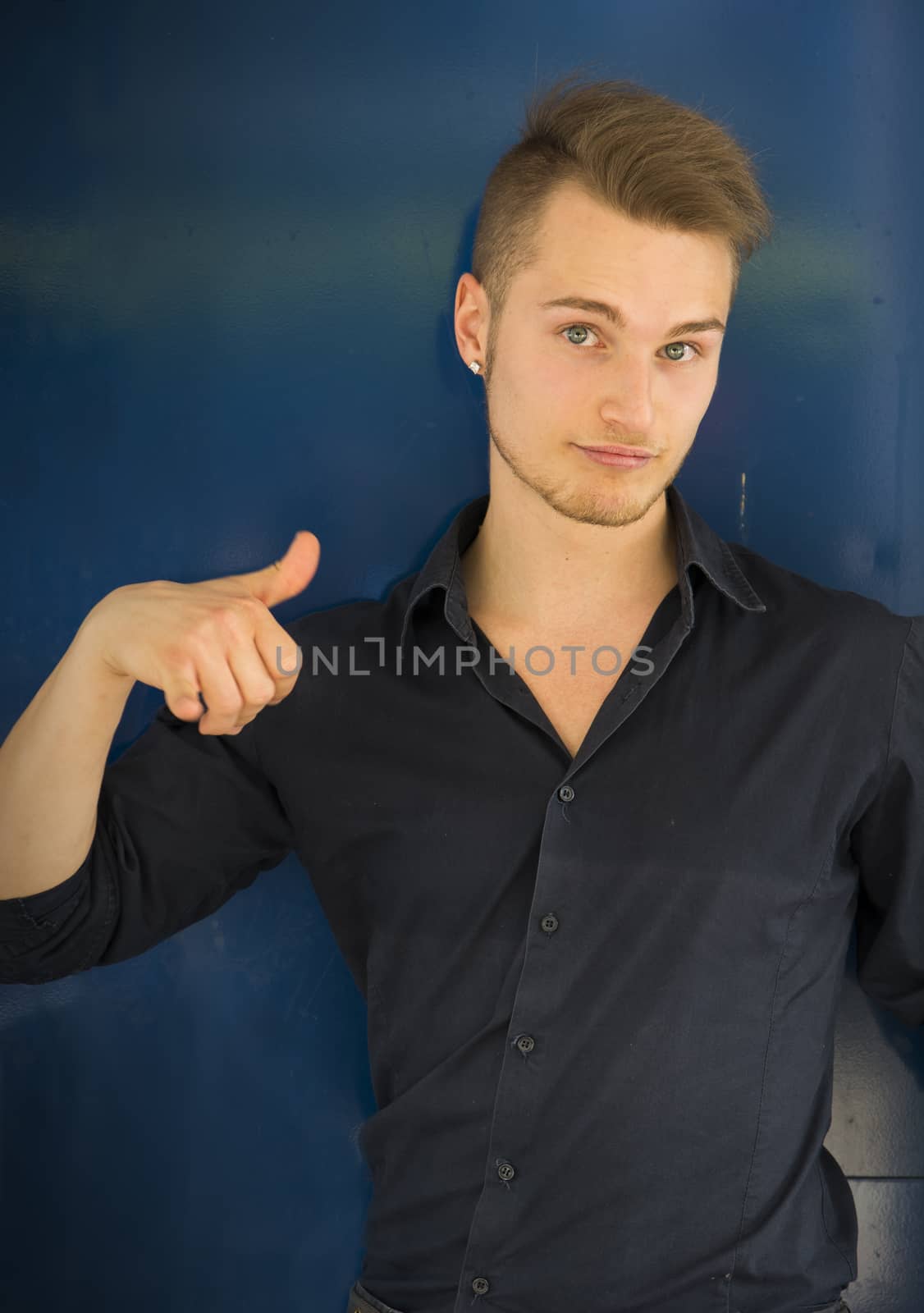 Attractive blond, blue eyed young man smiling and doing thumb up sign with one hand. Blue background