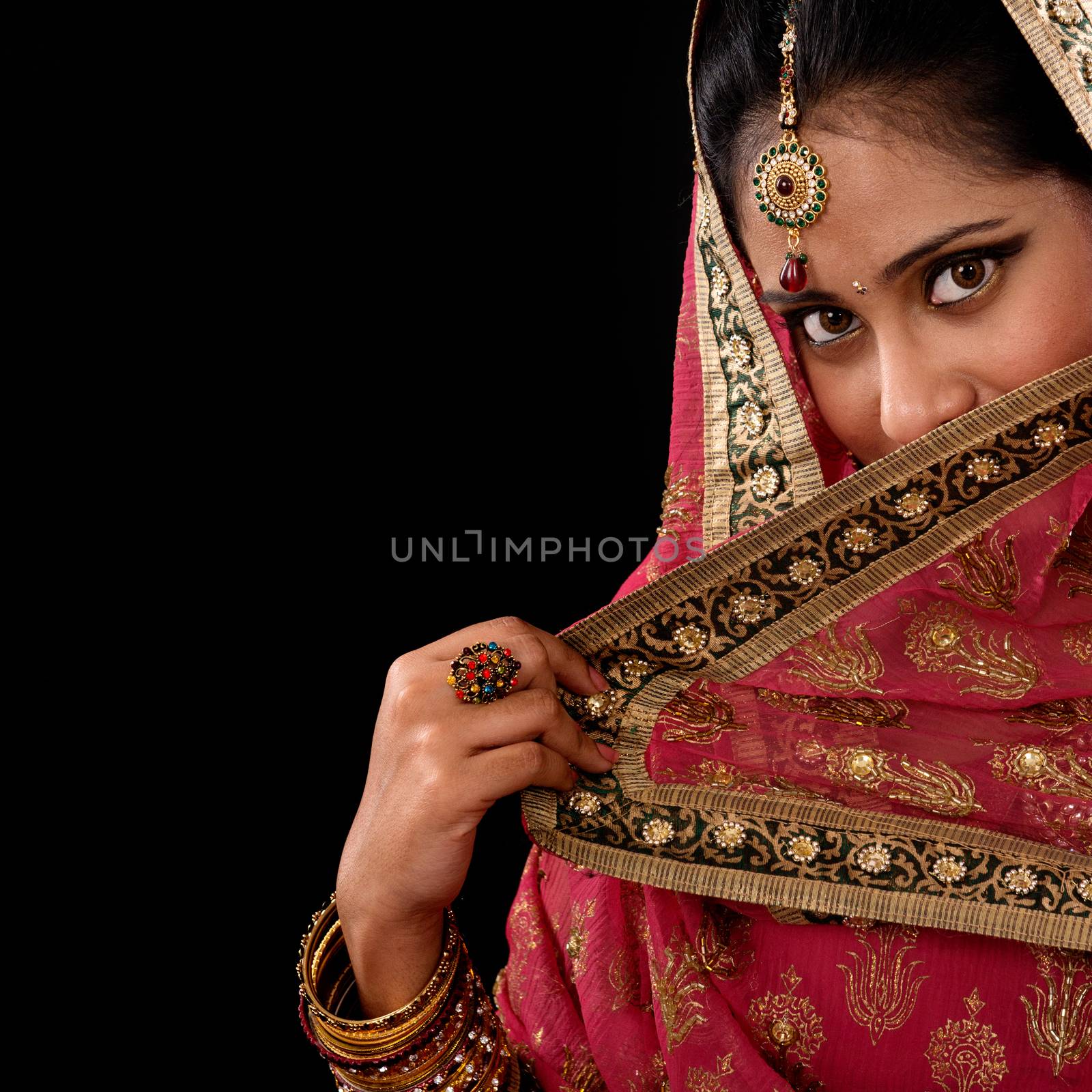 Portrait of beautiful mystery young Indian woman covering her face by veil, looking at camera, copy space at side, isolated on black background.
