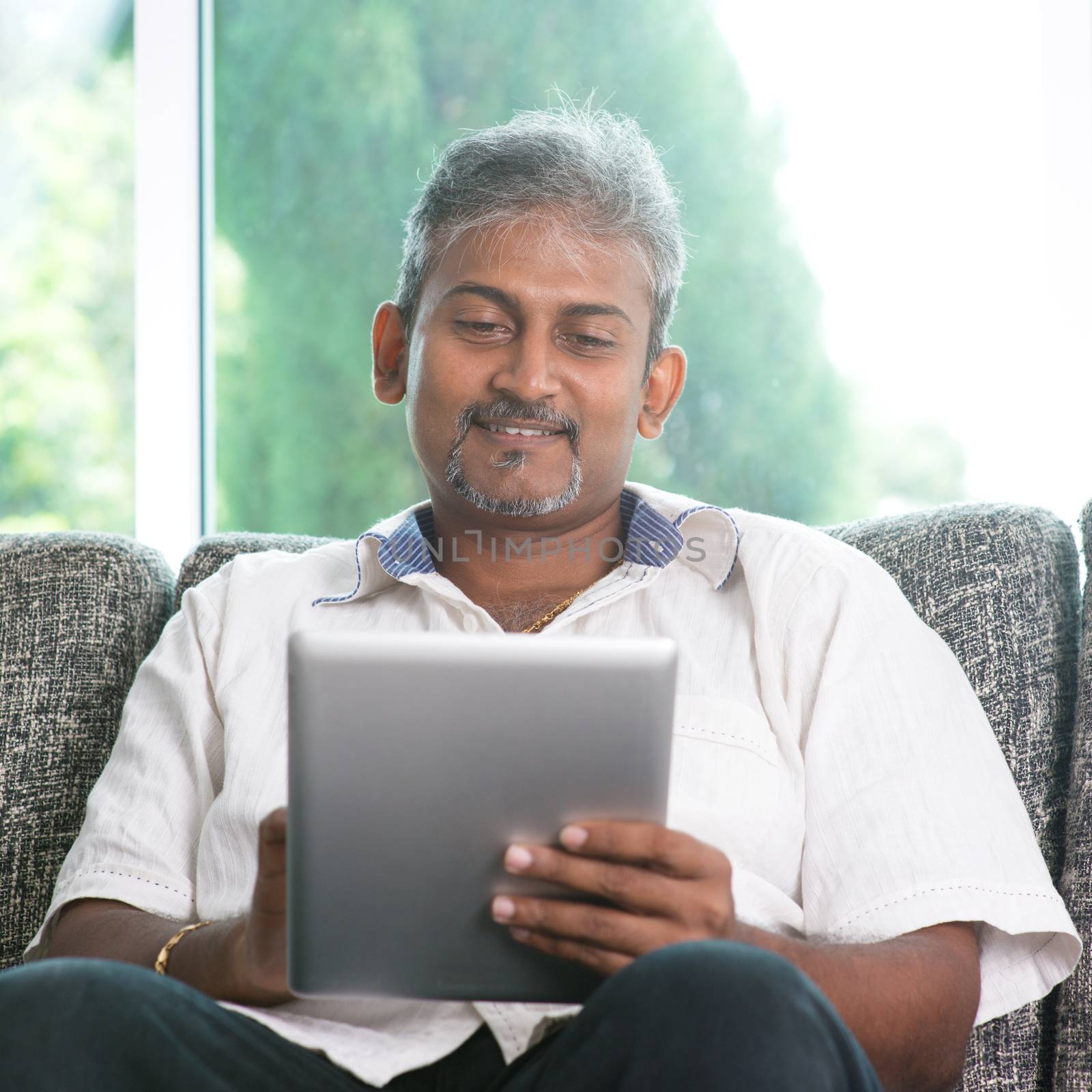 Portrait of middle aged Indian man reading on digital tablet computer and smiling at home.