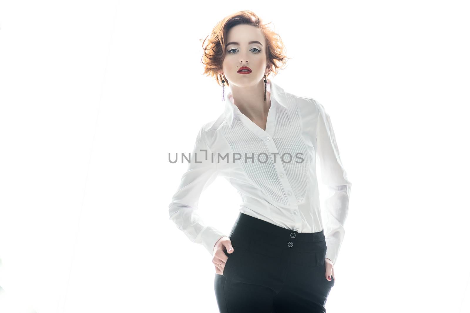 the beautiful stylish red-haired girl with red lipstick in a white blouse looks directly, holding hands on hips