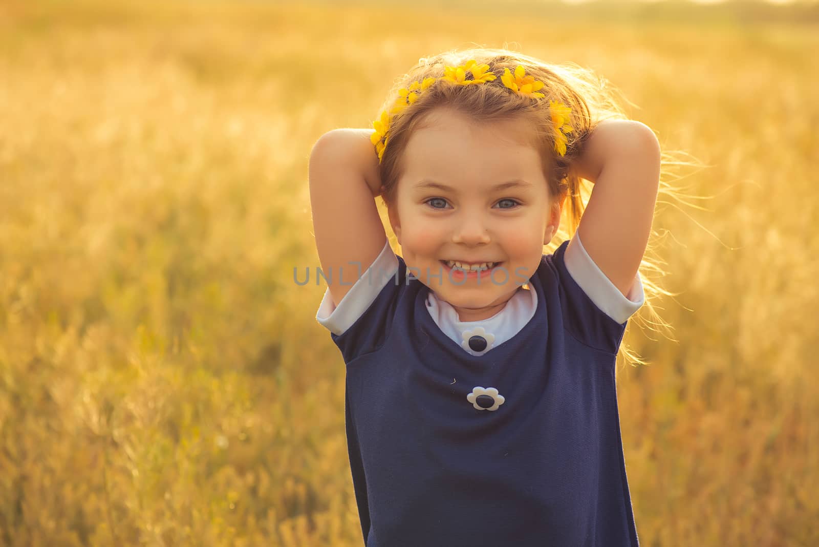the beautiful little girl in a blue dress against a field laughs, holds hands behind the head by odindia