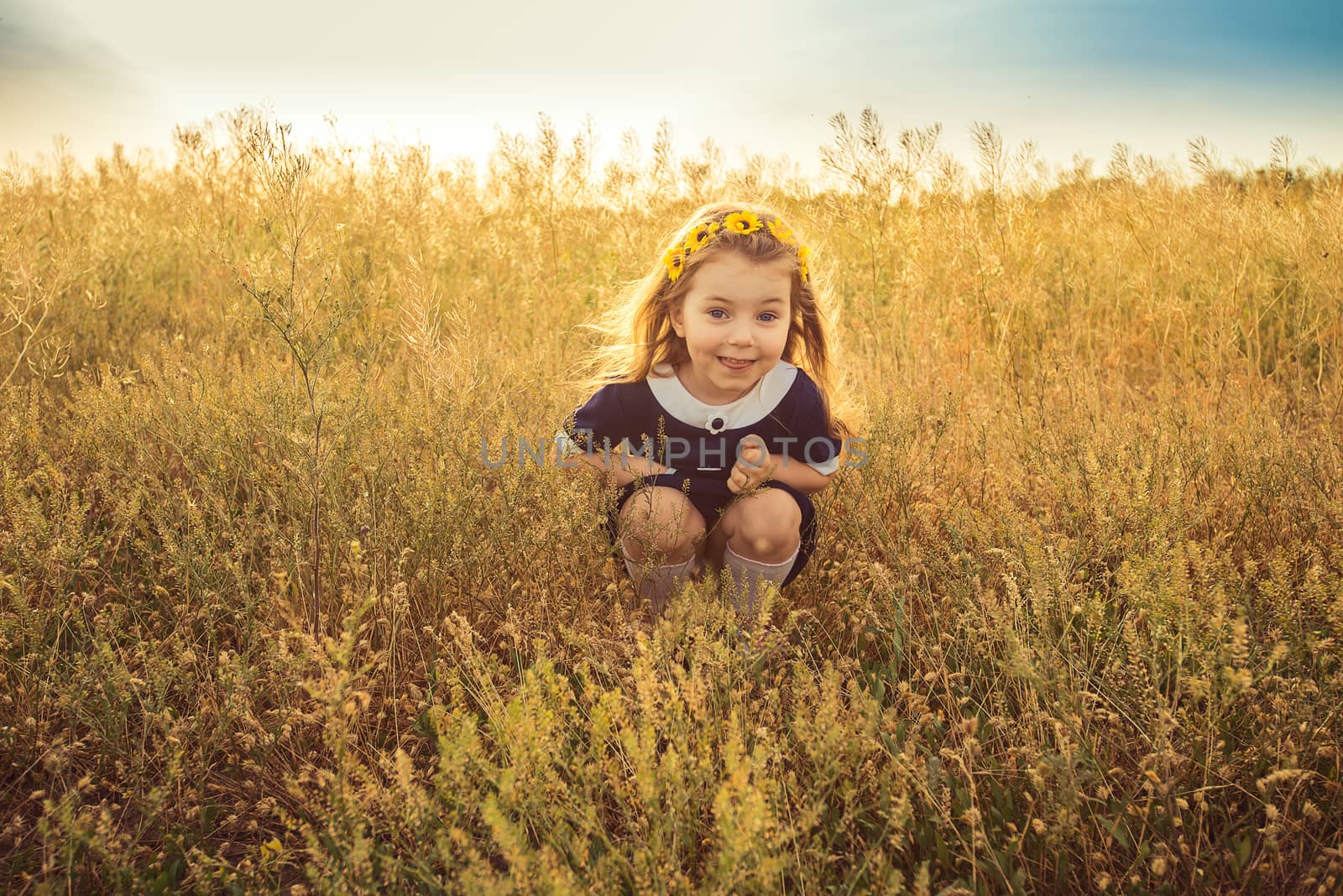 the beautiful little girl in a blue dress sitting in the field smiles by odindia