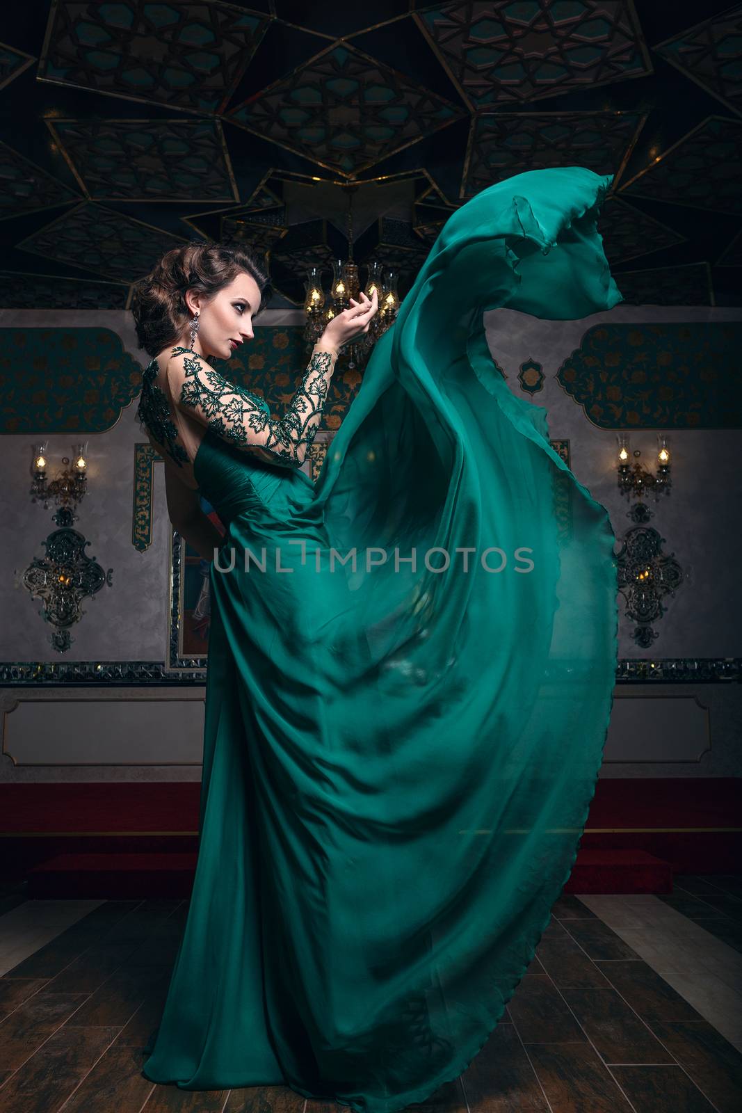 Beautiful woman in a green long dress on a background of richly decorated room. Fashion woman in fluttering dress.