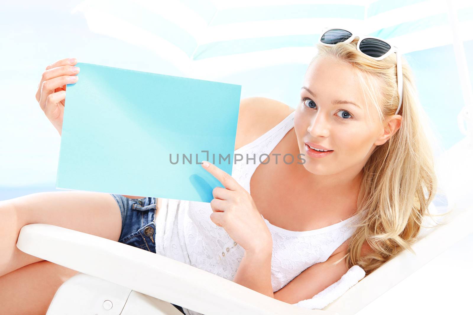 Beautiful blonde woman sitting on a lounger beach holding a blue sign with space for text.