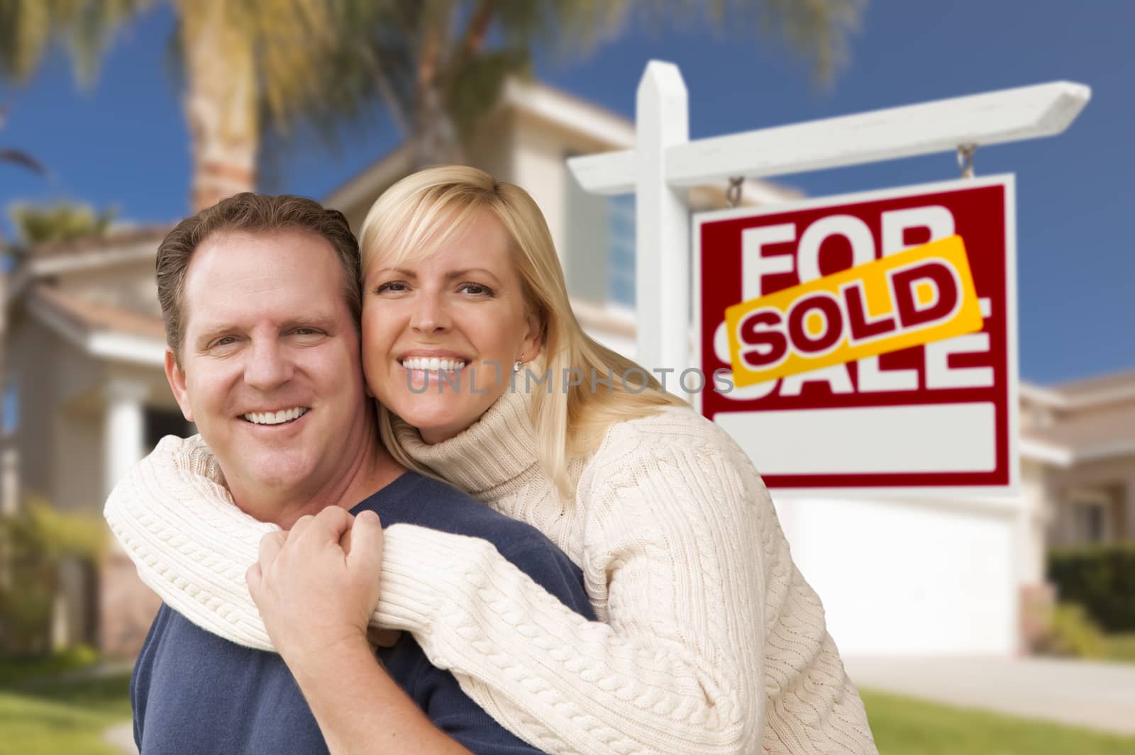 Happy Couple Hugging in Front of Sold Real Estate Sign and House.