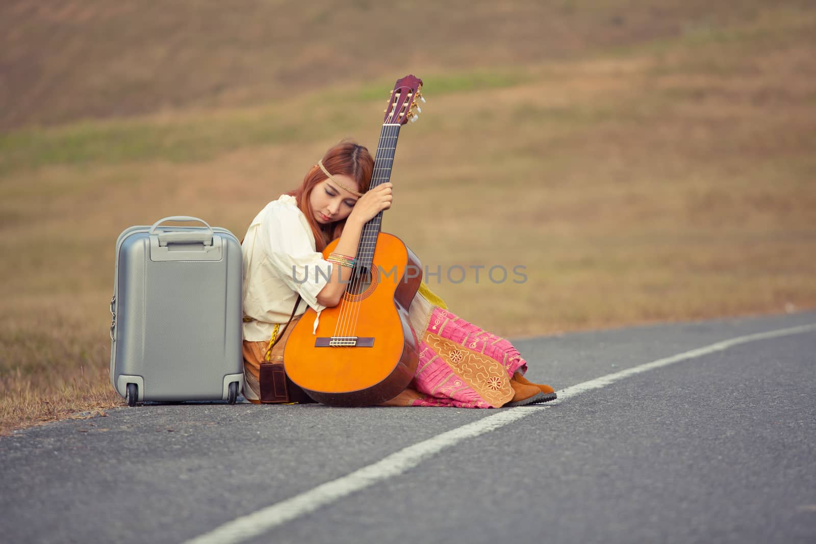 Young hippie woman with guitar and backpack sitting on a countryside road