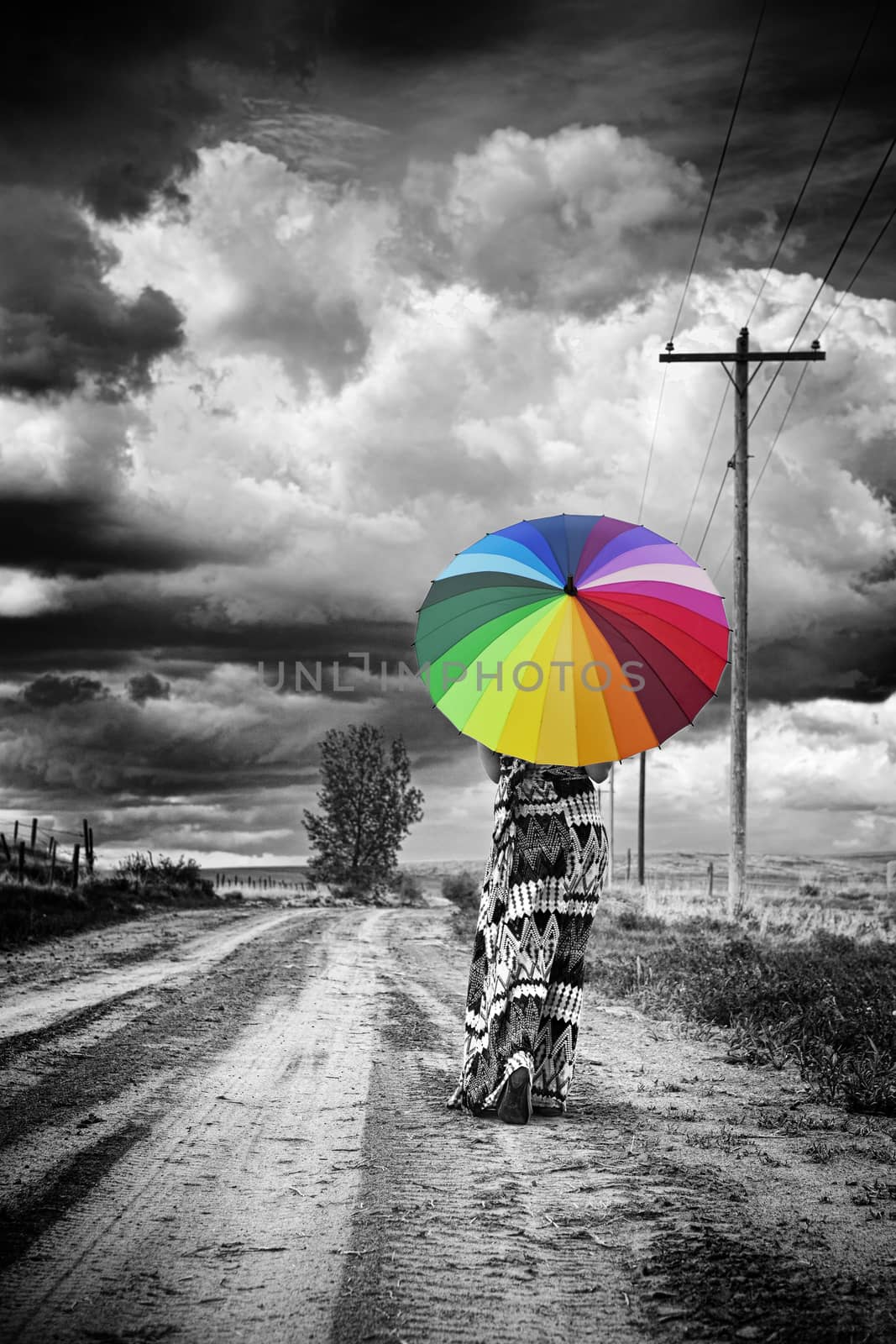 A woman walks alone along, an old dirt road with ominous clouds above.  Gritty black and white with selective color.