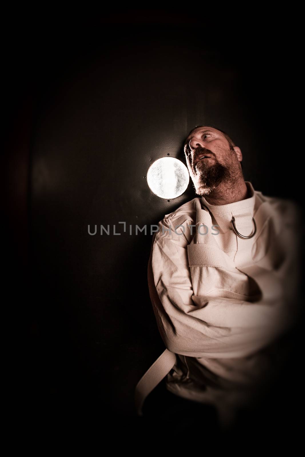 Photo of an insane man in his forties wearing a straitjacket standing in a cell of an asylum with the light from the hallway streaming in.