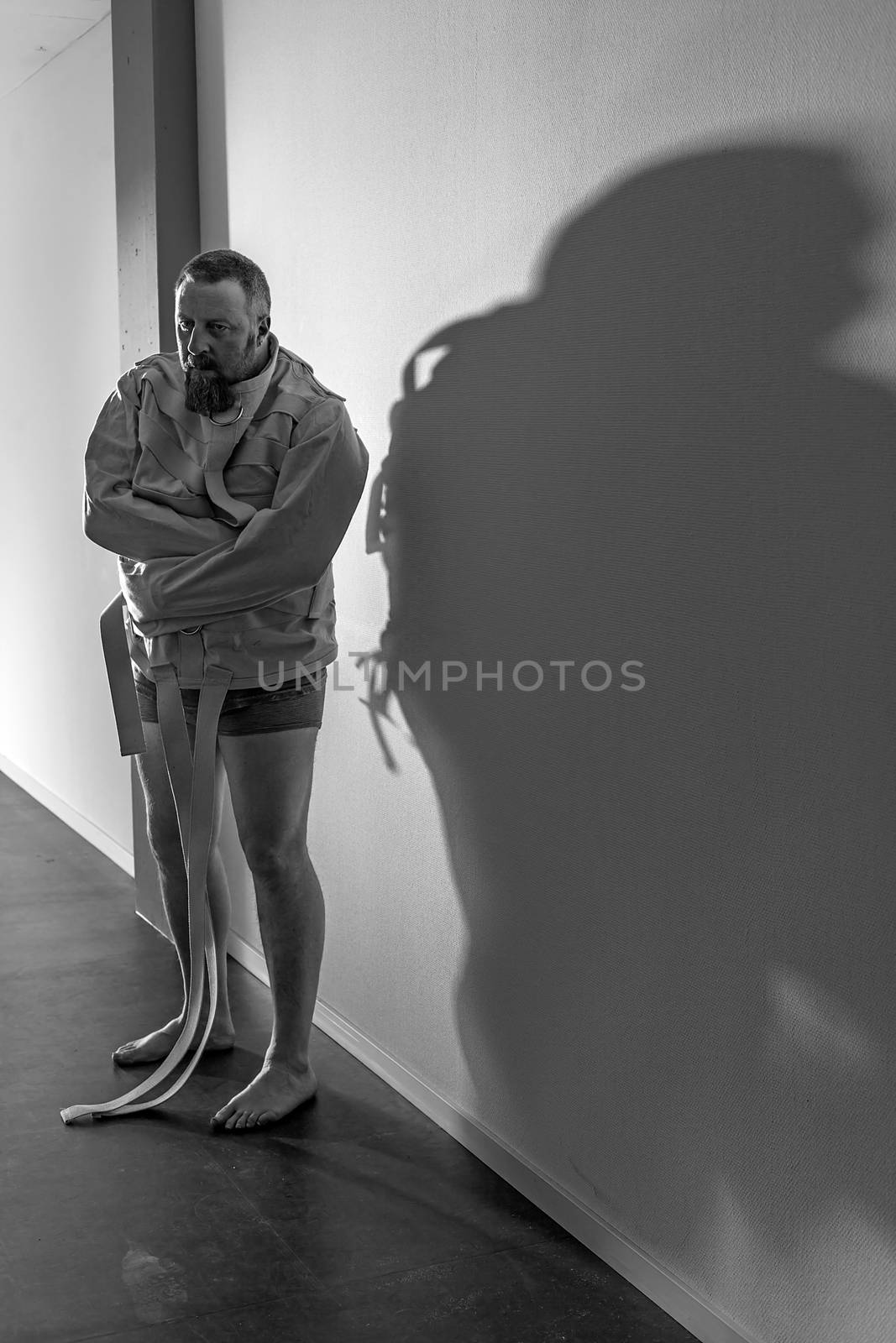 Photo of an insane man in his forties wearing a straitjacket standing in a hallway of an asylum.