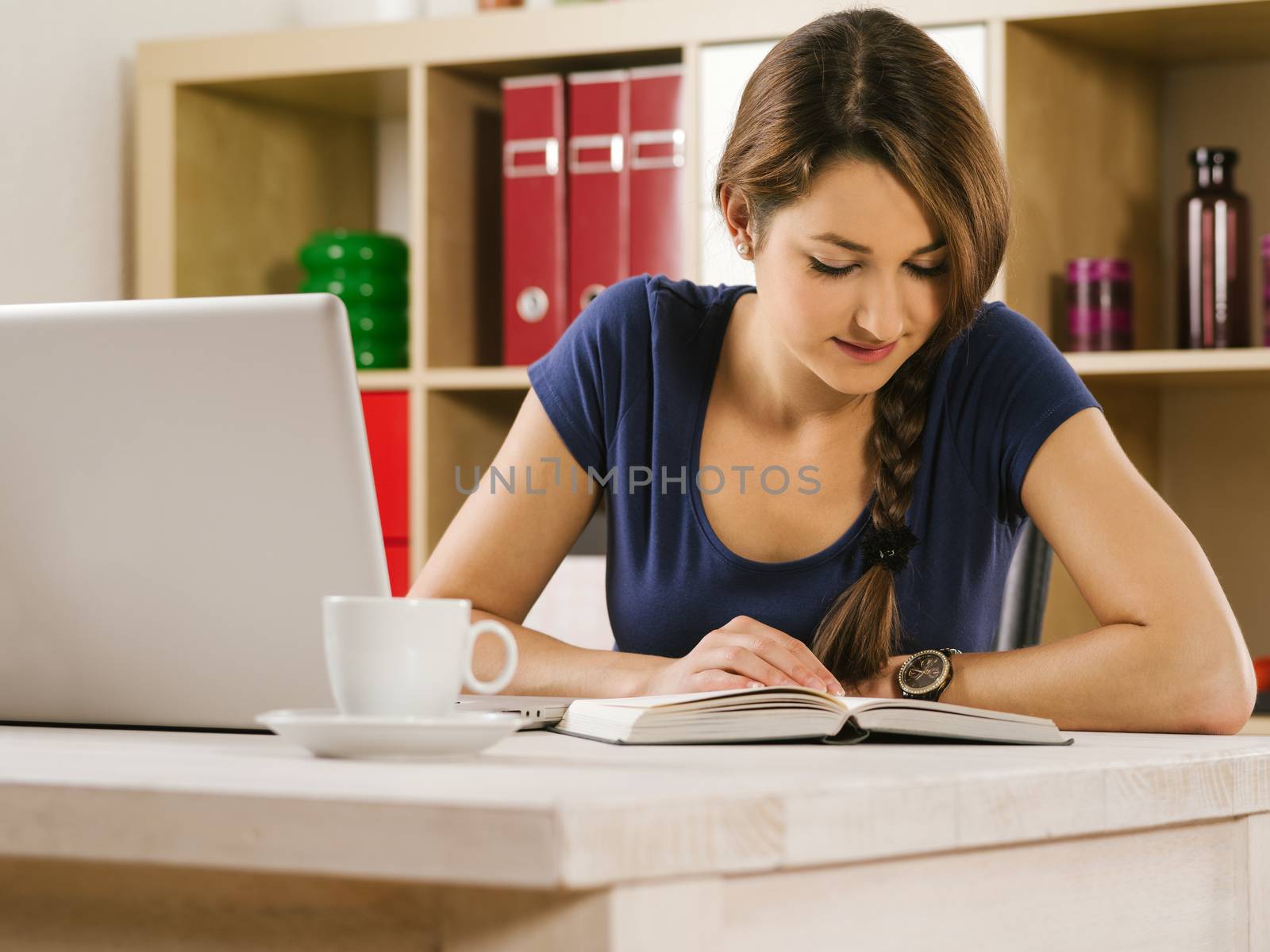 Photo of a beautiful woman using a laptop, drinking coffee, and reading a book at home or an office.
