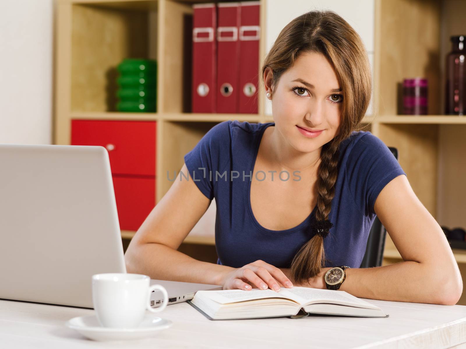 Photo of a beautiful woman using a laptop, drinking coffee, and reading a book at home or an office.