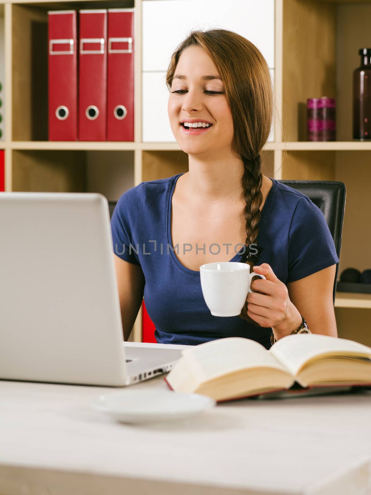 Photo of a beautiful laughing woman using a laptop and drinking coffee at home or at her office.