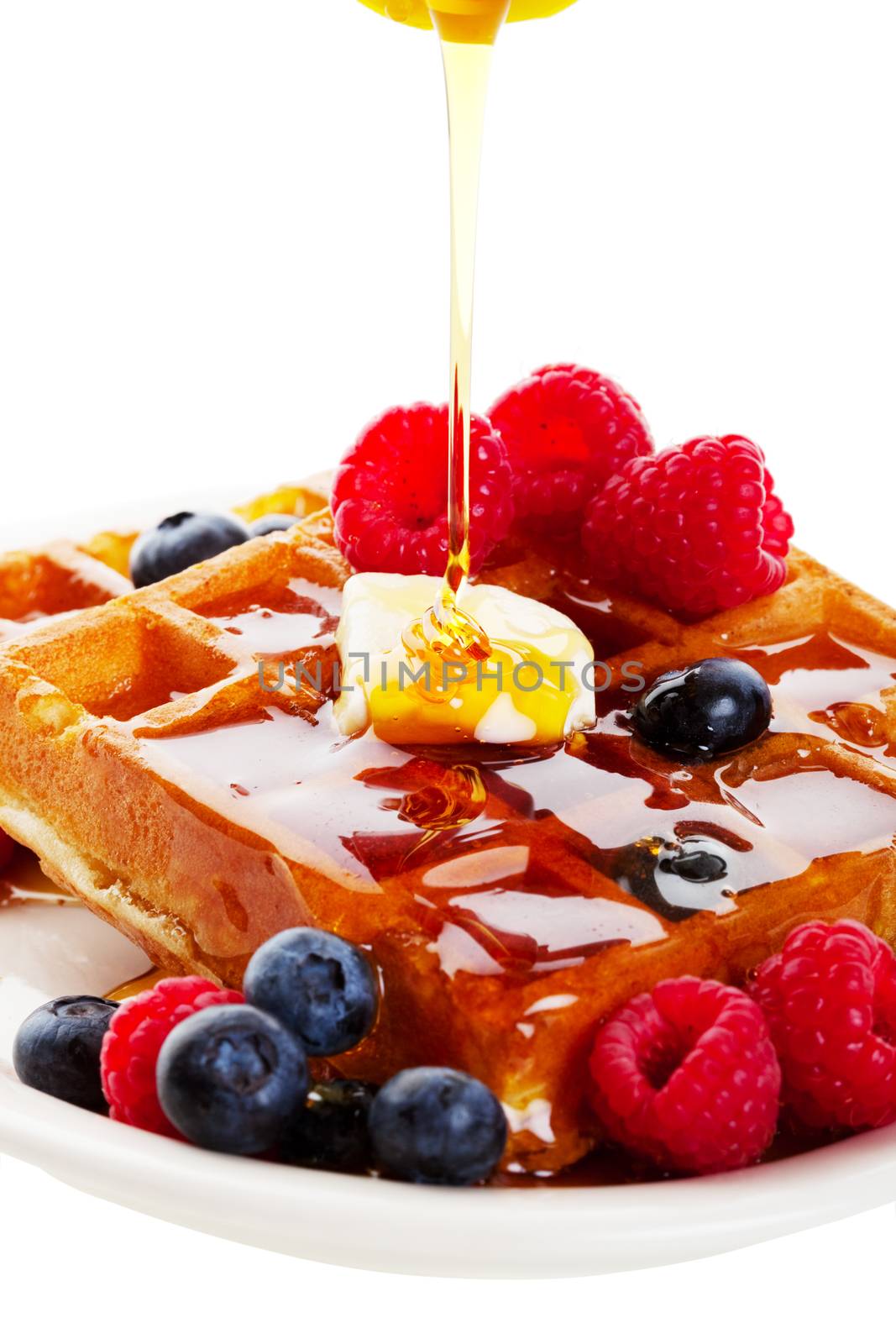 A stream of golden Canadian maple syrup adds the finishing touch to a delicious breakfast of belgian waffles with fresh raspberries and blueberries.  Shot on white background.
