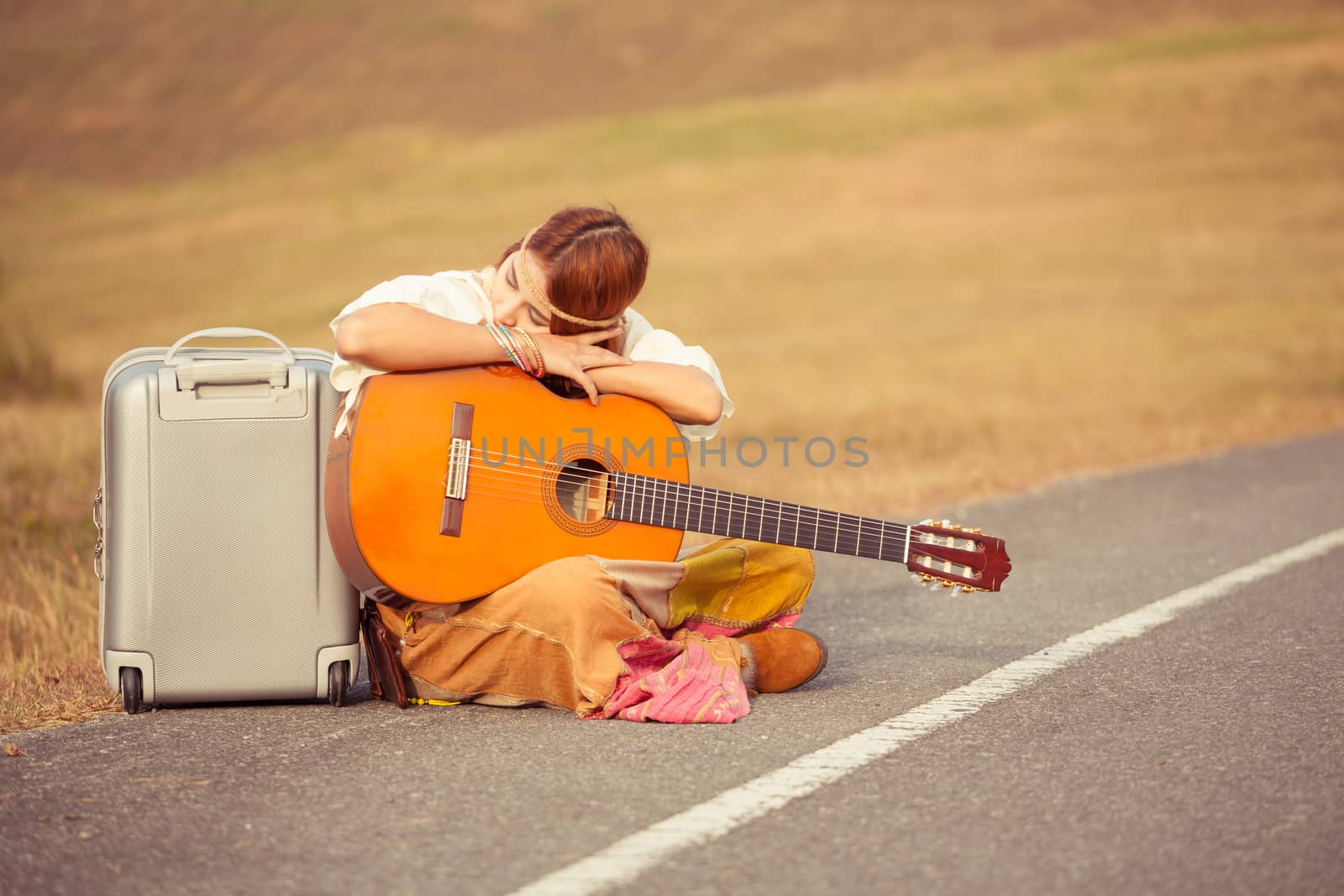 Young hippie woman with guitar and backpack sitting on a countryside road