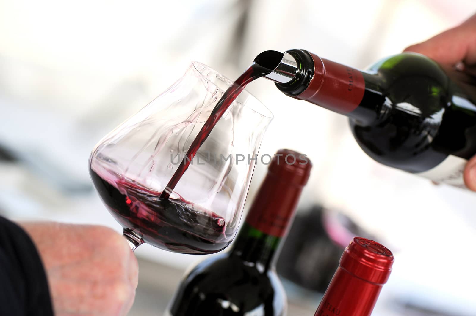 Tasting wine in a vinery-A man's hand is pouring red wine into a glass with a corkscrew set down. Lot of wine bottles in the background with shadows.