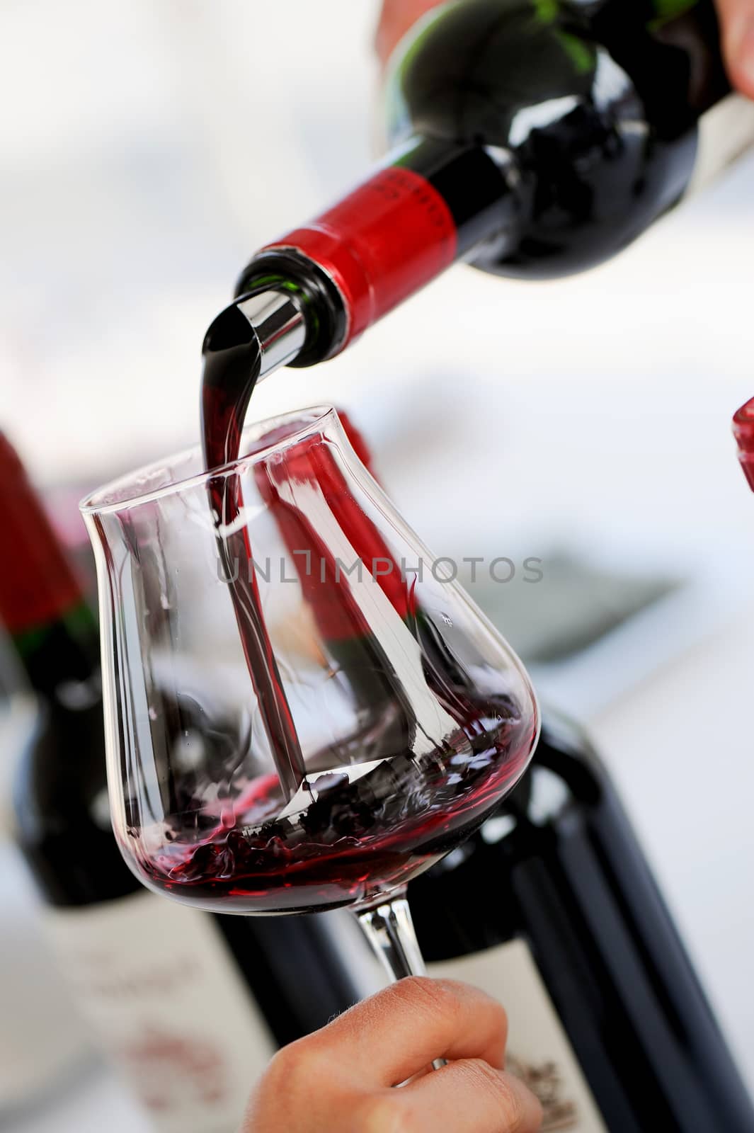 Tasting wine in a vinery-A man's hand is pouring red wine into a glass with a corkscrew set down. Lot of wine bottles in the background with shadows.