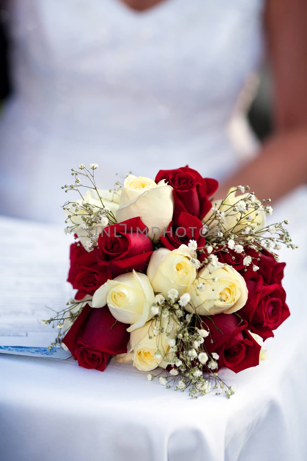A wedding bouquet of red and white roses with wisps of babybreath, stands in the forefront while a bride signs in the muted background.