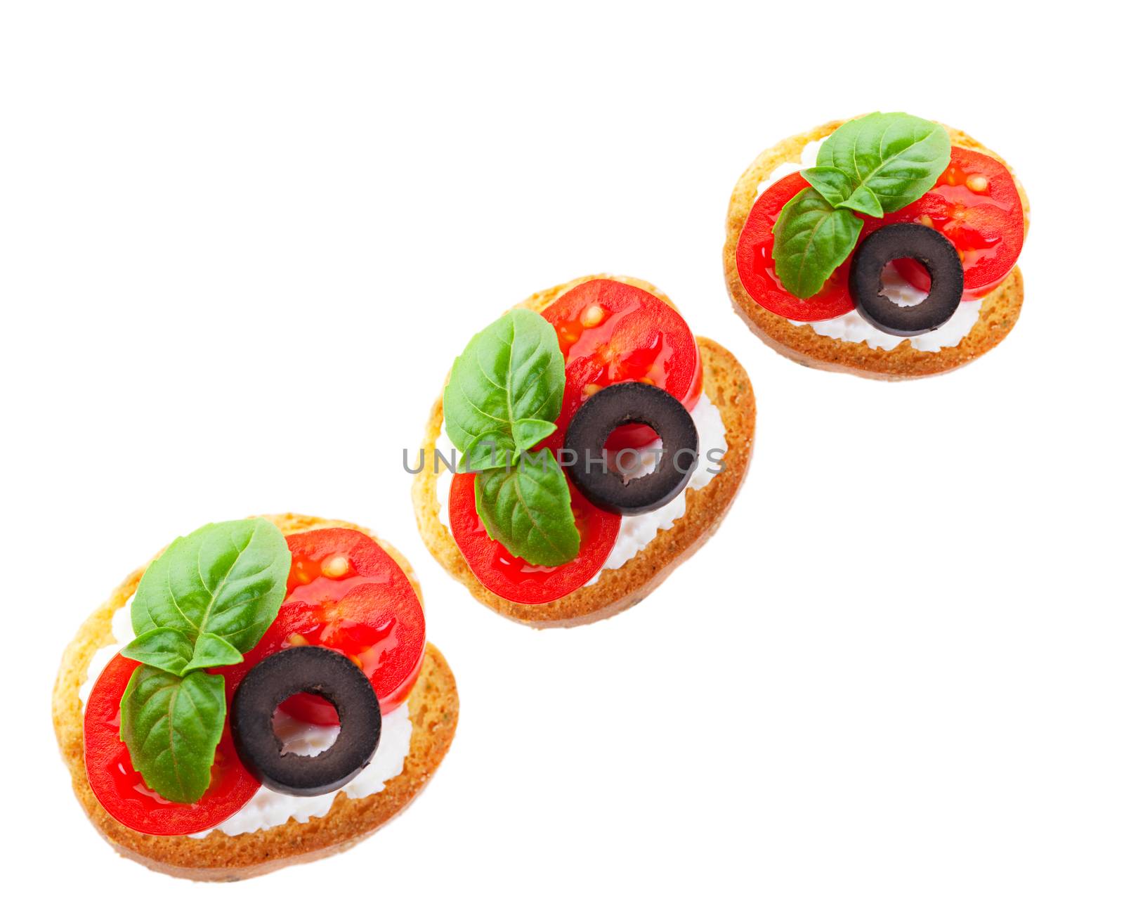 Juicy slices of baby tomato on a goat cheese covered baguette cracker, garnished with a slice of black olive and a fresh sprig of basil.  Shot on white background.