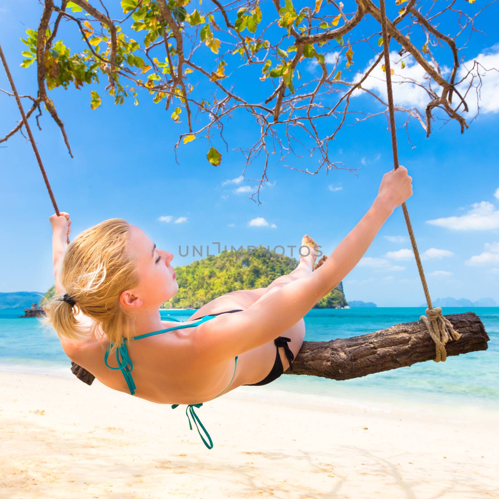 Lady swinging on the picture perfect tropical beach with the view of the island and turquoise coral reef on a sunny summer day.