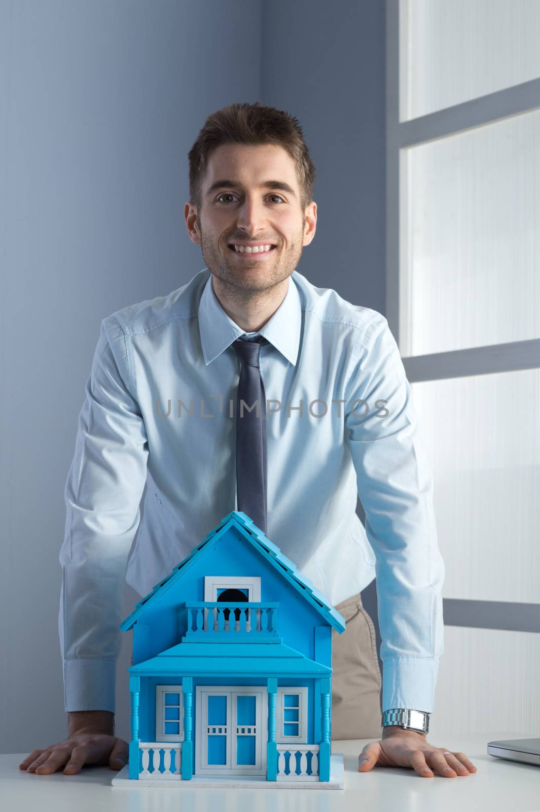 Young real estate agent leaning on desk with model house.